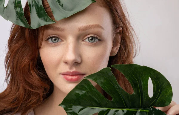 Vermelho de cabelos jovens do sexo feminino posando com folhas verdes Monstera, fundo cinza — Fotografia de Stock