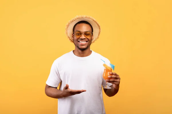 Millennial negro chico mostrando vaso de delicioso cóctel tropical en amarillo fondo del estudio — Foto de Stock