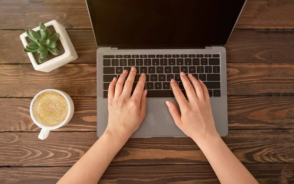 Mãos de mulheres no teclado de computador portátil, senhora que trabalha no computador com xícara de café e pequena fábrica no pote na mesa de madeira — Fotografia de Stock