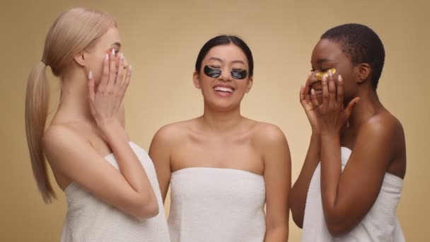 Позитивні різноманітні жінки, загорнуті в рушники, кладуть гелеві плями під очима і сміються, бежевий студійний фон — стокове відео