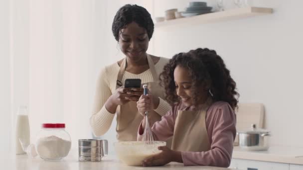 가족 요리 블로그. 아프리카 계 미국인 어머니가 반죽을 만들고 있는 딸 과 스마트폰으로 비디오를 녹화하는 모습 — 비디오