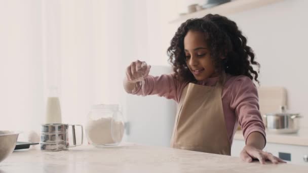 Gadis afrika kecil yang lucu mengenakan celemek menuangkan tepung ke atas meja sebelum berlutut adonan, memasak di dapur — Stok Video