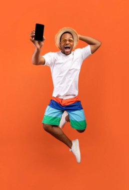 Heyecanlı genç zenci adam canlı yayında cep telefonunu turuncu arka planda siyah boş ekranla gösteriyor.