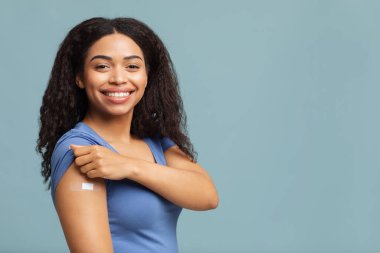 Covid-19 aşımı yaptırdım. Mutlu Afro-Amerikalı kadın aşı olmuş kolunu gösteriyor antiviral enjeksiyondan sonra, mavi arka plan