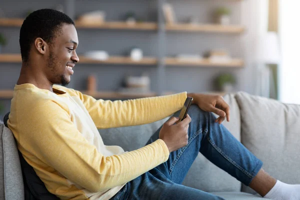 Sonriente chico negro reclinado en el sofá, utilizando el teléfono inteligente —  Fotos de Stock
