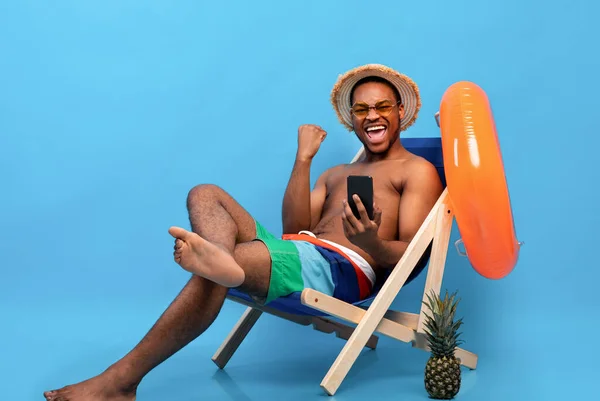 Funky svart kille i badkläder håller smartphone, känsla upphetsad, sitter i solstol, gester JA, blå bakgrund — Stockfoto