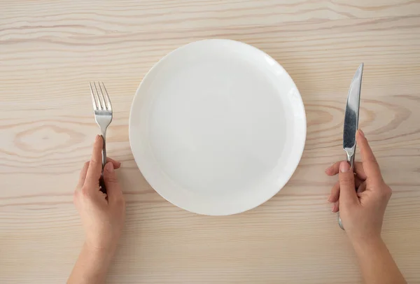 Placa blanca vacía y manos femeninas sosteniendo tenedor de plata y cuchillo sobre fondo de mesa de madera, vista superior —  Fotos de Stock