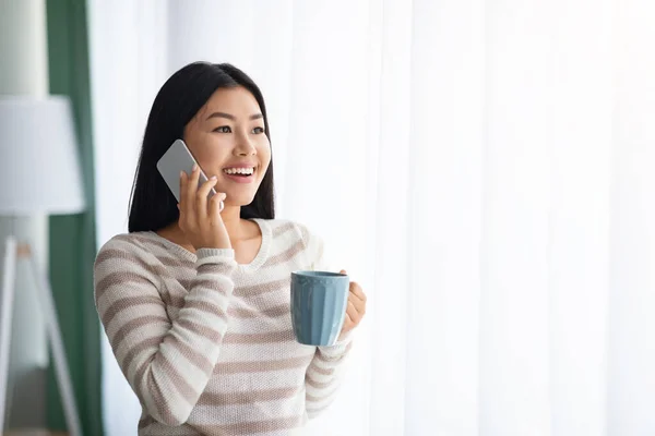 Mooie jonge aziatische vrouw drinken koffie en praten op mobiele telefoon in de buurt van venster — Stockfoto
