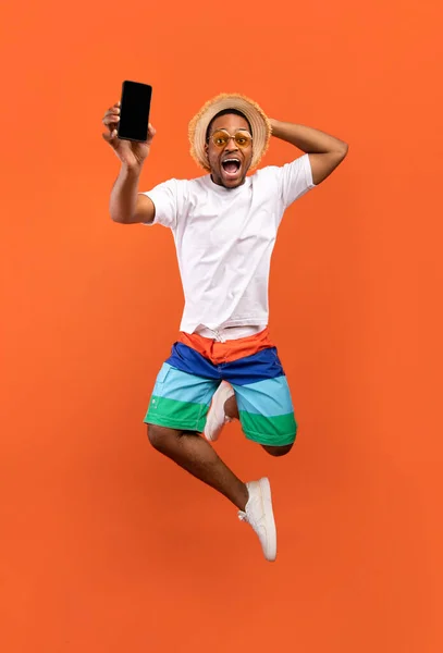 Excited young black guy jumping on air, showing cellphone with black empty screen on orange background, full length — 스톡 사진