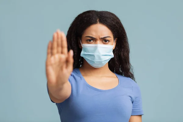 Sociaal afstandconcept. Ernstige Afro-Amerikaanse dame met beschermende masker tonen stop gebaar tegen blauwe achtergrond — Stockfoto
