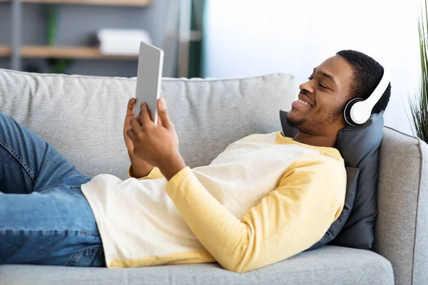 Smiling black guy laying on couch, watching movies on pad — 스톡 사진
