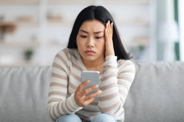 Haberler kötü. Üzgün Asyalı Kadın Tutucu Akıllı Telefon, Endişeli Ekrana Bakıyor