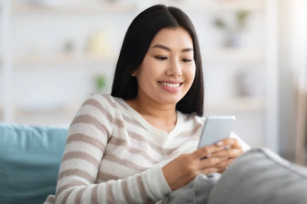 Fröhliche junge Asiatin sendet Nachrichten auf dem Smartphone, während sie sich zu Hause entspannt — Stockfoto