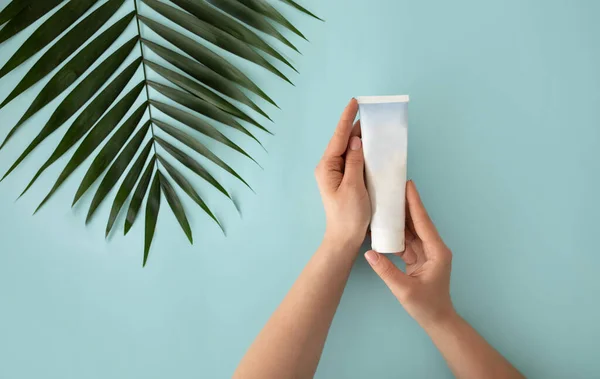 Mãos femininas segurando tubo branco para creme, hidratante, loção ou limpador facial em fundo azul com folha tropical — Fotografia de Stock