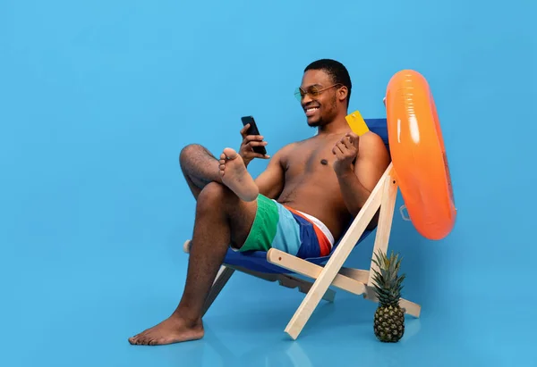 Hombre negro sonriente usando tarjeta de crédito y teléfono inteligente para ir de compras en línea mientras se enfría en el sillón sobre fondo azul —  Fotos de Stock