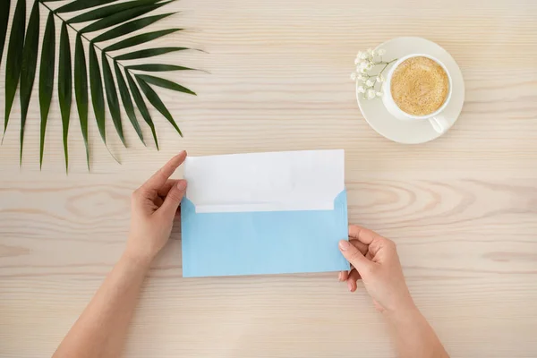 Draufsicht der sich öffnenden weiblichen Hände Grußkarte mit blauem Umschlag auf dem Tisch mit Tasse Kaffee und tropischem Blatt — Stockfoto