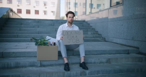 Crise de emprego. Chateado jovem gerente sentado na escada com suas coisas pessoais e segurando cartaz Procurando um emprego — Vídeo de Stock