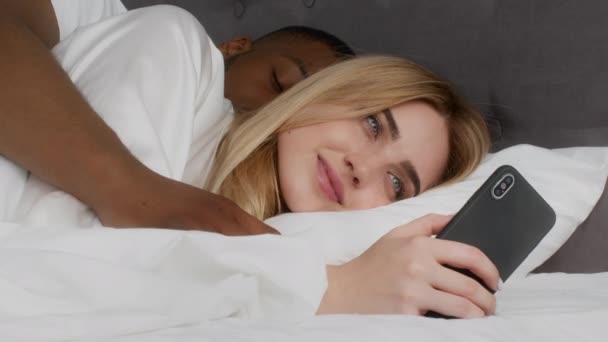 Jovencita acostada en la cama y en red en el teléfono inteligente, esposo afroamericano durmiendo y abrazándola — Vídeo de stock