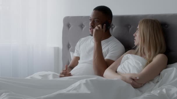 Crisis de relaciones en la cama. Ocupado afroamericano hombre hablando en el teléfono inteligente, señora molesta sentirse disgustado — Vídeos de Stock