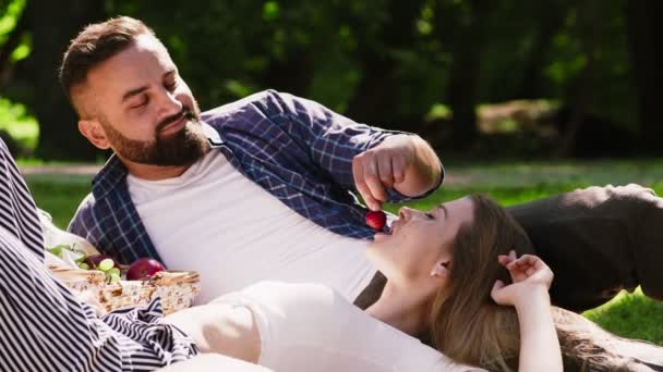 Almoço romântico no parque. Feliz casal afetuoso desfrutando de piquenique no parque verde, homem alimentando mulher com frutas frescas do verão — Vídeo de Stock