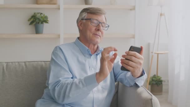 Hymyilevä vanhempi mies silmälaseissa istuu sohvalla ja käyttää matkapuhelinta kotona, juttelemassa perheensä kanssa — kuvapankkivideo