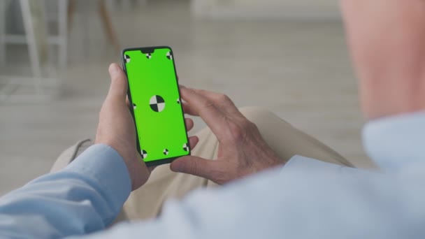 Smartphone moderno con pantalla de croma verde en manos del hombre mayor que se relaja en el sofá en casa, sobre la vista del hombro — Vídeos de Stock