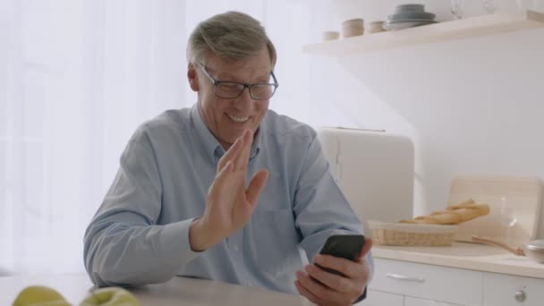 Vanhempi mies, joka käyttää älypuhelinta videopuheluun kaukaisen perheensä kanssa, istuu keittiössä, heiluttaen kädestä puhelimeen — kuvapankkivideo