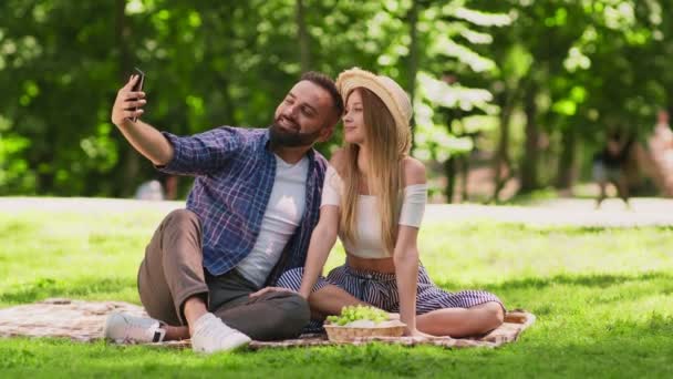 Счастливая кавказская пара влюблена, делая селфи вместе, позируя на камеру смартфона, сидя на одеяле в парке — стоковое видео