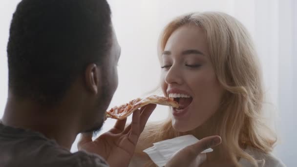 Разнообразная пара, поедающая пиццу, кормящая друг друга флиртом в помещении — стоковое видео