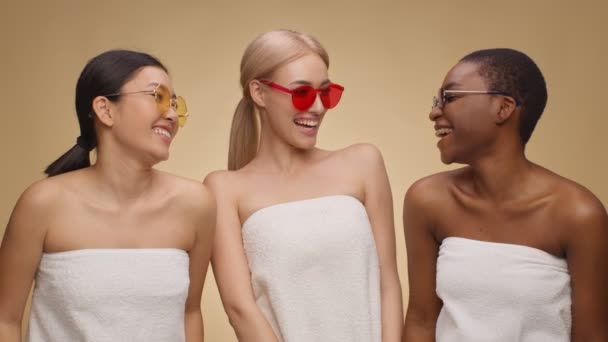 Tre glade multietniske damer indpakket i håndklæder og iført solbriller ser på hinanden og griner – Stock-video