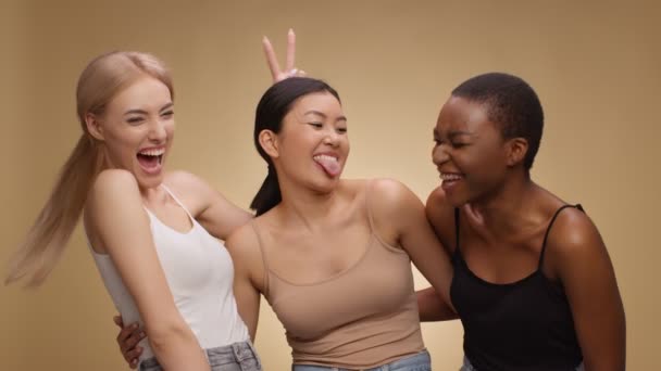 Tres felices amigos multiétnicos riendo juntos, haciendo muecas y sacando lenguas, fondo beige, cámara lenta — Vídeos de Stock