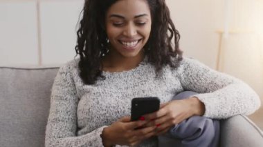 Genç mutlu kıvırcık saçlı Afro-Amerikan bayan akıllı telefondan mesaj atıyor, sosyal medyada arkadaşlarıyla mesajlaşıyor ve gülüyor.