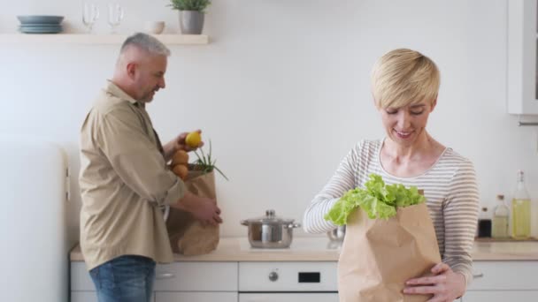 Pareja feliz desembalaje bolsas después de compras de comestibles de pie en la cocina — Vídeos de Stock