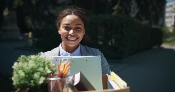 Happy african american woman manager carrying box with personal belongings and smiling widely to camera outdoors — Stock Video