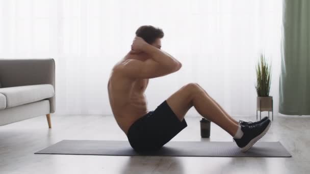 Vue latérale de jeune athlète masculin musclé pratiquant l'entraînement abdominal à la maison, faisant des craquements sur le sol, au ralenti — Video