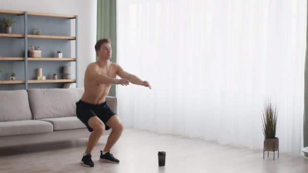 Young man with naked torso doing squats at home interior, practicing fitness workout alone at living room, tracking shot — Stock Video