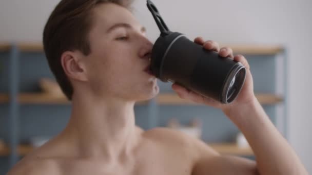 Lächelnder junger hemdloser Bodybuilder, der Protein oder Wasser aus der Sportflasche trinkt und zu Hause lächelt, Porträt aus nächster Nähe — Stockvideo