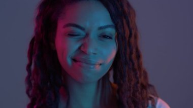 Flört havasında. Sevimli Afro-Amerikalı kadın kameraya gülümsüyor, mor neon ışıklarla poz veriyor, ağır çekimde.