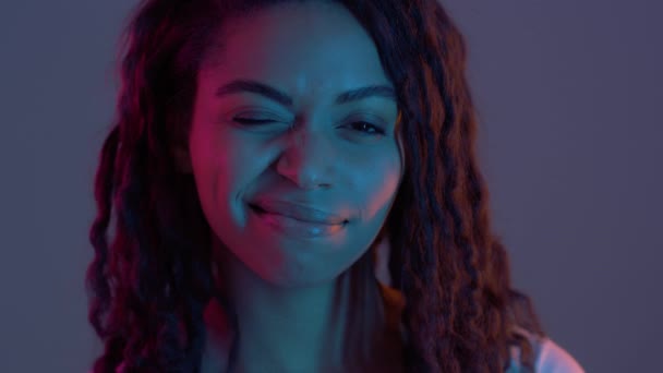 Humeur pétillante. Jolie dame afro-américaine ludique clignant des yeux et souriant à la caméra, posant dans des néons violets, au ralenti — Video