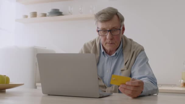 Zakupy online przez Internet. Nowoczesny senior korzystający z karty kredytowej i laptopa do zakupów internetowych, siedzi w kuchni — Wideo stockowe