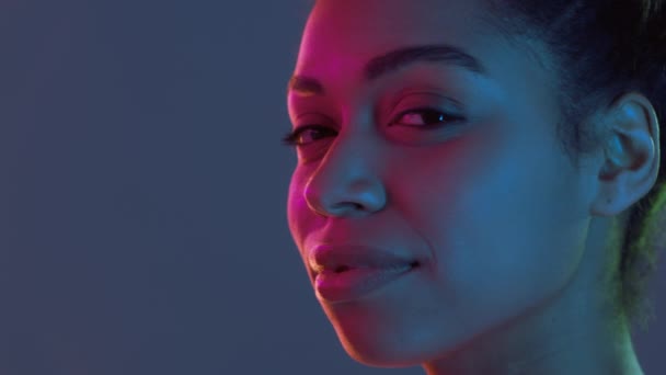 Profil portret młodej afrykańskiej kobiety pozytywnej obracając się twarzą w twarz do kamery i uśmiechnięte, neonowe światła tło — Wideo stockowe