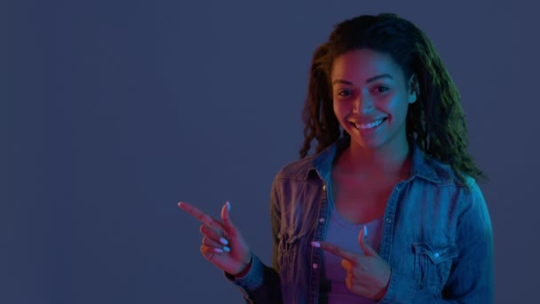 Νεαρή θετική αφροαμερικανή γυναίκα που δείχνει τα δάχτυλα στην άκρη, προσδιορίζοντας κενό χώρο για μακιγιάρισμα, ποζάροντας σε φώτα νέον — Αρχείο Βίντεο