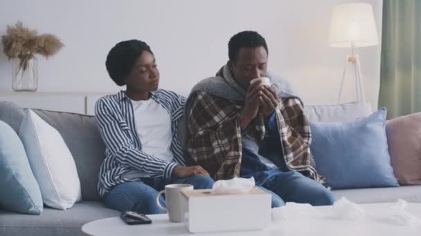 Assistenza alla famiglia. Giovane uomo afroamericano malato che beve tè curativo, la sua amorevole moglie che lo abbraccia e lo sostiene — Video Stock