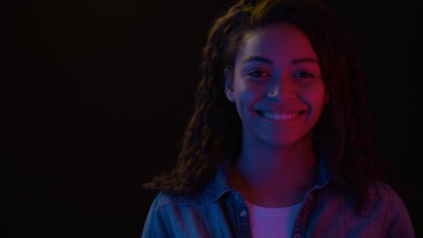 Young cheerful african american woman smiling to camera in neon lights, black background with empty space — Stock Video