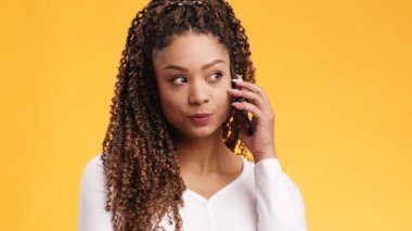 Afro saçlı, cep telefonuyla arkadaşlarıyla konuşan ve gülümseyen neşeli Afro-Amerikan kadının stüdyo fotoğrafı.