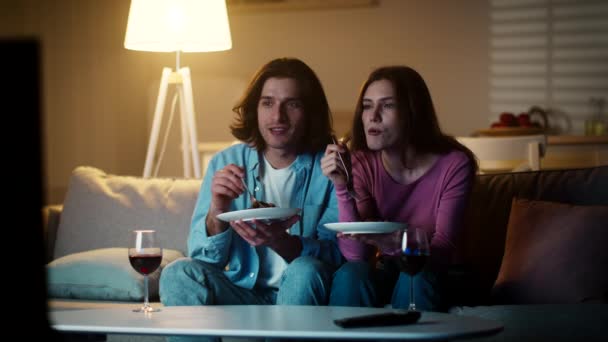 La vida cotidiana de la familia joven. Los cónyuges positivos disfrutan de una sabrosa cena con vino tinto y viendo el programa de televisión por la noche — Vídeo de stock