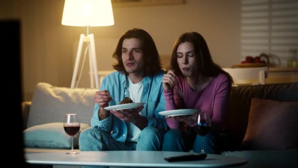 Молодой человек и женщина смотрят реалити-шоу по телевизору, обсуждают его и ужинают с вином дома вечером — стоковое видео