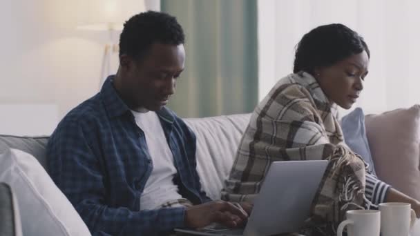 Jonge zwarte man werkt op laptop thuis, zijn zieke vrouw niezen en snuit neus, zitten verpakt in ruitjes op de bank — Stockvideo