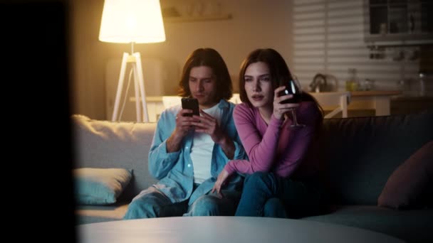 Молода байдужа пара проводить вечір вдома, жінка дивиться телевізор і п'є вино, чоловік спілкується на смартфоні — стокове відео