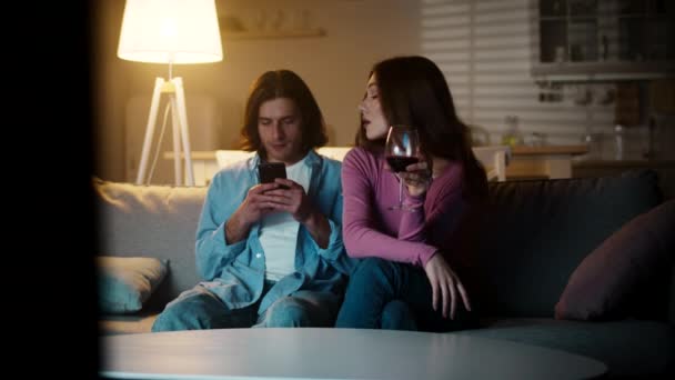 Ung kvinna tittar på tv med vin, ringer henne spela på smartphone pojkvän att följa med henne, visar film på kvällen — Stockvideo